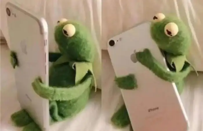 科米蛙專注地躺臥手持iPhone，手機背面印有「iPhone」字樣，此形象源自布偶秀。 科米蛙,Kermit,iPhone,手機,專注,布偶秀,Muppets,青蛙,玩手機,躺