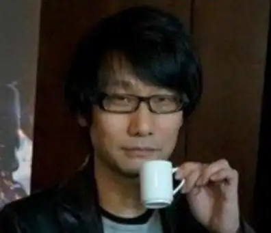 戴眼鏡的黑髮男子穿深色外套，手持白色小杯，面露沉靜且微帶笑意，置身於模糊的室內環境。為知名電玩製作人小島秀夫。 小島秀夫,電玩製作人,遊戲設計師,戴眼鏡男子,喝咖啡,沉靜,微笑